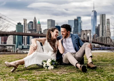 Braut und Bräutigam sitzen auf einer Wiese vor einer modernen Skyline während eines Hochzeitsfotoshootings