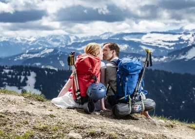 Braut und Bräutigam mit Wanderrucksäcken sitzen auf einem Berg und küssen sich mit Alpenpanorama im Hintergrund
