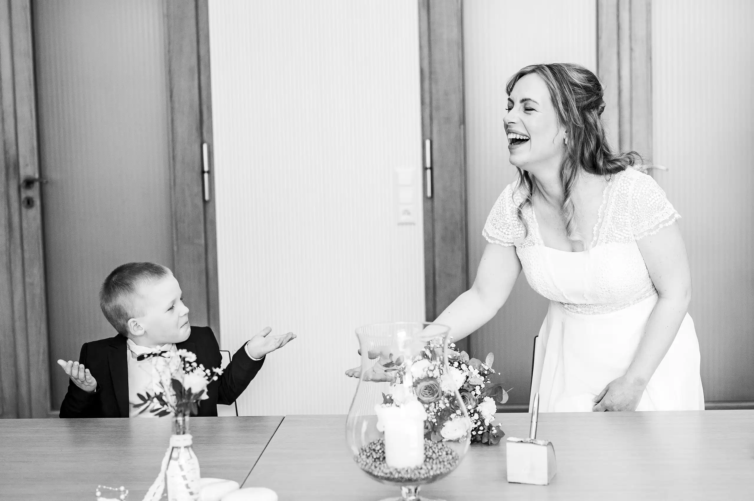 Hochzeitsfotografie – Emotionale Momente bei der Trauung Braut lacht während ein Junge im Anzug neben ihr bei der Trauung am Tisch sitzt