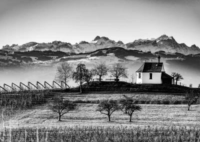 Kleine Kapelle auf Hügel vor verschneitem Alpenpanorama Schwarz-Weiß